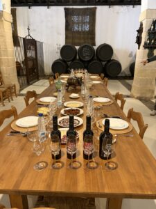Long wooden table set for a meal with wine bottles, water, glasses, and plates; rustic room with stacked wine barrels in the back.
