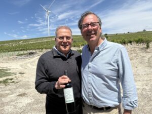 Two men pose for a selfie in a vineyard, with a wind turbine in the blue sky background; one holds a bottle of red wine.
