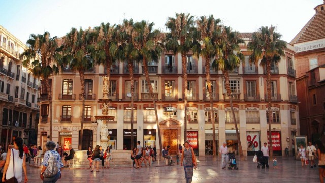 People walking on the main square of Malaga, Plaza de la Constitucion. It was was the main square in Malaga since the middle ages. Under Arab rule, it was called Square of the Four Streets. In 1812, the plaza got its present name. A renaissance style marble fountain made in Genoa called the Fuente de Genova is located at one end of the Plaza. Courtesy image Plaza de la Constitucion in Malaga