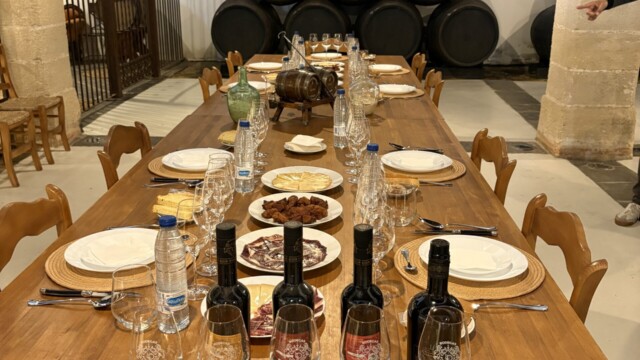 Long wooden table set for a meal with wine bottles, water, glasses, and plates; rustic room with stacked wine barrels in the back.