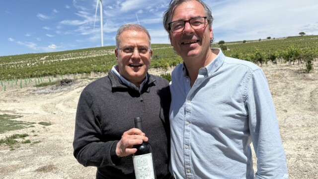 Two men pose for a selfie in a vineyard, with a wind turbine in the blue sky background; one holds a bottle of red wine.